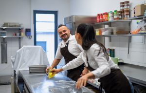 A chef and cook cleaning the workspace after doing dishes indoors in restaurant kitchen.